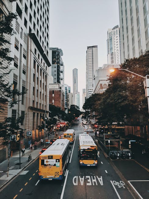 A view of a busy urban street in daytime, lined with tall modern office buildings and commercial properties on both sides. Several yellow buses are traveling along the road, with one bus in the foreground displaying route and service information. Pedestrians are visible walking on the pavement, some near parked bikes and motorcycle parking areas. Streetlights and traffic signs are present, and trees line parts of the sidewalk, adding greenery to the scene. The lighting suggests overcast weather, and the overall environment reflects a typical cityscape suitable for home relocation or furniture transport, as part of a professional moving service, such as those provided by Man with Van Leyton, operating in the Leyton area.