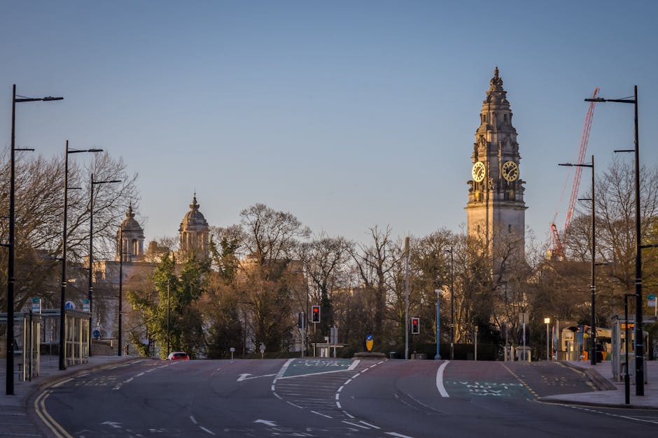 A view of a city street with a tall historic clock tower in the background, part of a larger building complex with domed structures. In the foreground, an empty road curves gently to the right, with v