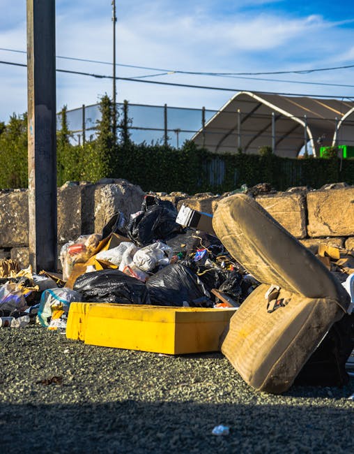 An outdoor scene showing a pile of discarded household waste including black and transparent plastic bin bags, a yellow plastic storage container, and a large old beige upholstered armchair with visible dirt and wear. The waste is situated on a gravel surface next to a stone wall and near a metal pole with overhead power lines. In the background, there are green bushes, a fenced area, and a partially visible large tent or industrial structure under a partly cloudy blue sky. The image relates to waste removal and clutter clearance, which key services offered by Man with Van Leyton for house removals and property clearance.