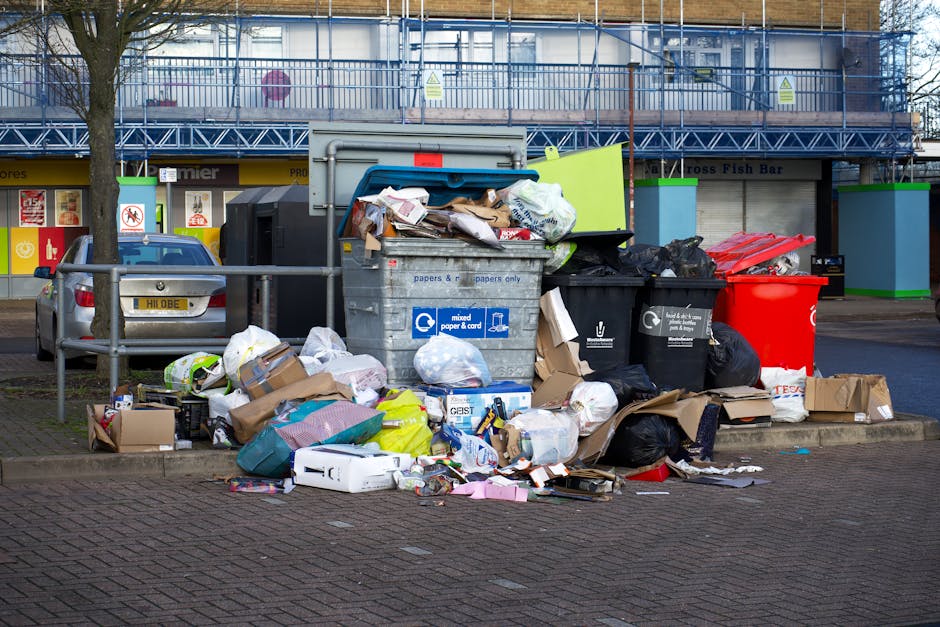 A large collection of overflowing waste and recycling bins located on a paved sidewalk area in front of a retail unit in Leyton, with cardboard boxes, plastic bags, paper, packaging materials, and discarded household items spilling onto the ground. One of the bins is open, revealing mixed paper and cardboard, while others are closed or tipped over. A red wheelie bin and black recycling bin are visible among the waste. Behind the bins, a street and storefronts with signage and a blue scaffolding structure are seen, indicating a commercial area. The waste is situated near parking spaces, with a parked silver car to the left and a metal railing separating the waste from the vehicle. The scene is captured in daylight, illustrating the typical urban street environment, which local residents and businesses might need assistance with during property clearances or removals, as provided by Man with Van Leyton.