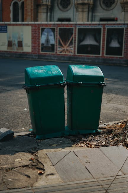 An outdoor scene showing a pile of discarded household waste including black and transparent plastic bin bags, a yellow plastic storage container, and a large old beige upholstered armchair with visible dirt and wear. The waste is situated on a gravel surface next to a stone wall and near a metal pole with overhead power lines. In the background, there are green bushes, a fenced area, and a partially visible large tent or industrial structure under a partly cloudy blue sky. The image relates to waste removal and clutter clearance, which key services offered by Man with Van Leyton for house removals and property clearance.