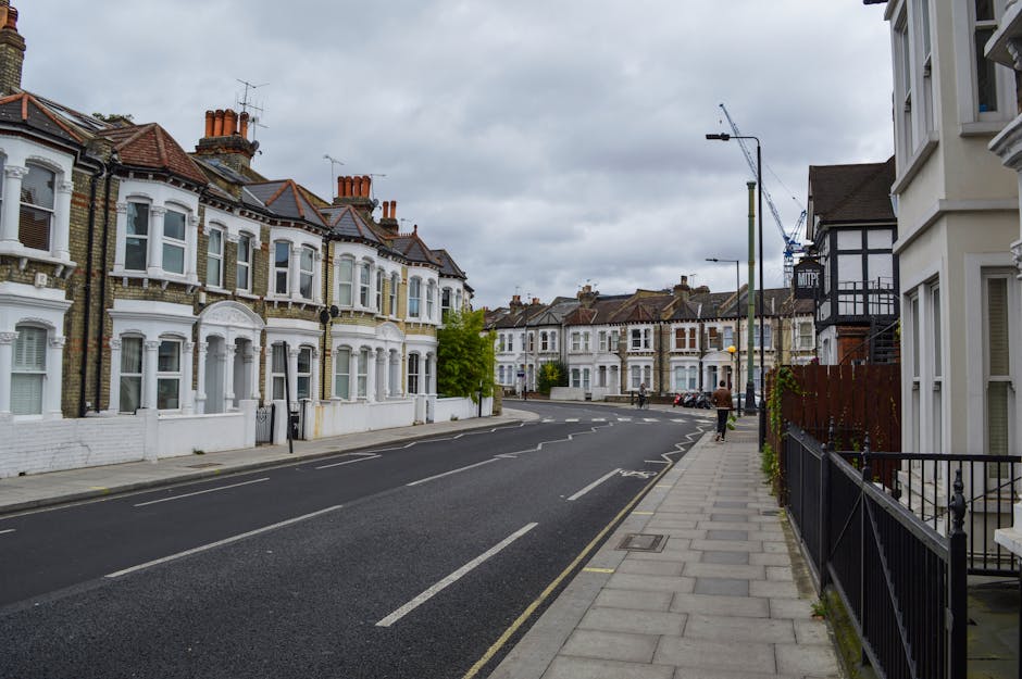 Photograph of a busy street in Leyton E10 with multiple parked and moving cars along the roadside, including sedans and hatchbacks, under clear blue skies with some white clouds. On the sidewalk, ther