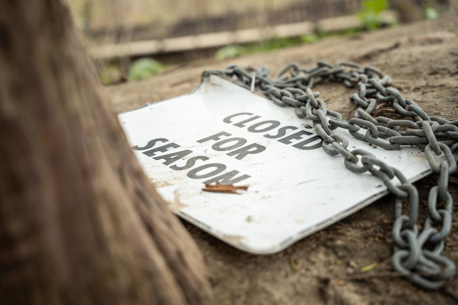 A close-up photograph of a weathered metal sign lying on the ground outside a property, with the words 'CLOSED FOR SEASON' visible on its surface. The sign is attached to a thick metal chain that is loosely draped across it, resting on a patch of dirt and small rocks near a tree trunk, with a wooden fence in the background. The setting appears to be an outdoor area, possibly part of a house or garden. The image highlights elements of property access restrictions, which can be relevant for property alterations or removals, and showcases workspace logistics involved in loading or clearing during house removals carried out by professionals like Man with Van Leyton.