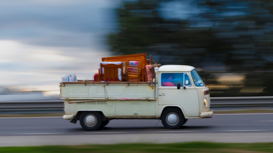 A white delivery van parked outdoors on a slight incline during dusk, with its side door open revealing an interior illuminated by warm light. Inside the van, wooden furniture, possibly chairs or smal