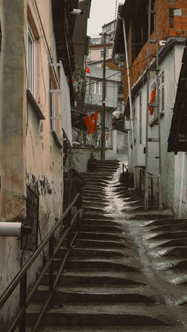 A narrow outdoor stairway made of concrete with uneven steps and metal handrails on both sides, situated between residential buildings in an urban area. The walls of the buildings are weathered, with visible signs of dirt and peeling paint. Loose debris and small puddles of water are present on the steps, indicating recent rain. At the top of the stairs, there are additional buildings with windows and balconies, some with hanging laundry and potted plants. The scene is lit by natural daylight, capturing a typical urban environment suitable for home relocation or furniture transport services, with Man with Van Leyton often assisting in such challenging access situations for part of their removals and moving logistics.