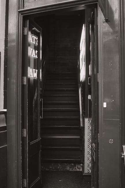 A black and white photograph of a narrow staircase viewed from the ground floor entryway of a property. The staircase has dark wooden steps with a slight sheen and is flanked by walls that appear to be painted or clad in dark material, creating a confined and enclosed space. The walls are plain, with minimal decoration, and there is a portion of a patterned tile visible at the bottom of the stairs near the threshold. The entrance to the staircase features a door frame without a door, leading directly into the stairwell. The lighting is muted, with some natural light coming from an upper window or opening, illuminating parts of the stairs and the surrounding walls. This setting is relevant to house removals and moving logistics, highlighting the challenge of navigating narrow staircases during furniture transport or home relocation, with the company Man with Van Leyton occasionally providing assistance in such situations.