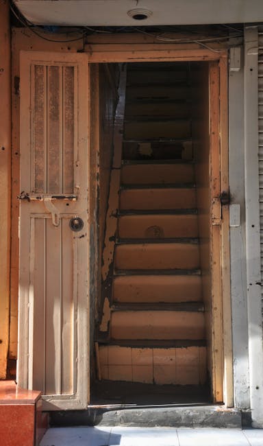 A narrow outdoor stairway made of concrete with uneven steps and metal handrails on both sides, situated between residential buildings in an urban area. The walls of the buildings are weathered, with visible signs of dirt and peeling paint. Loose debris and small puddles of water are present on the steps, indicating recent rain. At the top of the stairs, there are additional buildings with windows and balconies, some with hanging laundry and potted plants. The scene is lit by natural daylight, capturing a typical urban environment suitable for home relocation or furniture transport services, with Man with Van Leyton often assisting in such challenging access situations for part of their removals and moving logistics.