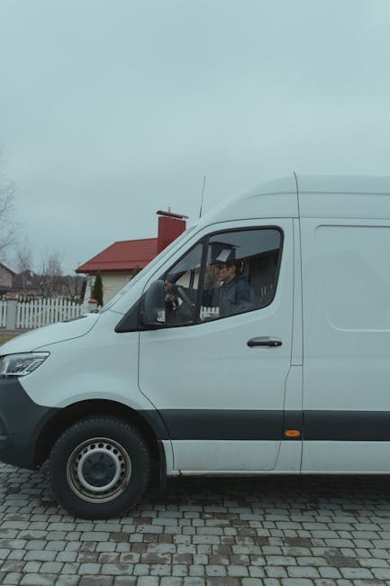 A white commercial van is parked on a cobblestone driveway in a residential area, with a man seated inside in the driver's seat. The van features a large side door and a visible front windshield, with
