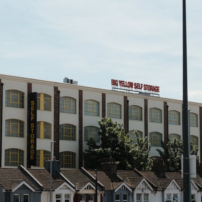 A multi-storey commercial building with a white facade and large arched windows outlined in yellow, featuring a vertical black sign reading 'SELF STORAGE' on the left side and a red sign on the roof displaying 'BIG YELLOW SELF STORAGE' in uppercase letters. Below the building, there are small residential houses with pitched roofs and chimney stacks. In front of the building, several green trees partially obscure the lower floors. The scene is taken during daytime with a partly cloudy sky, and a streetlamp is visible on the right side of the image. The setting illustrates the exterior of a self-storage facility, typical for house removals and packing services, with urban elements highlighting logistical considerations for home relocation or furniture transport by companies like Man with Van Leyton.