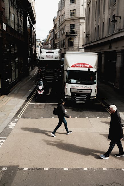 A street scene showing a moving van operated by Man with Van Leyton parked on a narrow urban road with tall, multi-storey buildings on either side. The white van has the 'uniserve' logo and website visible on its side and is positioned partially on the pavement, blocking part of the roadway. A small motorcycle is parked beside the van, and another vehicle is loaded behind it. In the foreground, a woman with dark hair, dressed in a black jacket and jeans, is crossing the street from left to right, while an older man with grey hair, wearing a dark coat, also crosses in the same direction. The scene depicts a typical home relocation process, with the loading area ready for furniture transport and packing materials likely inside the van, as part of a professional removals service in Leyton.