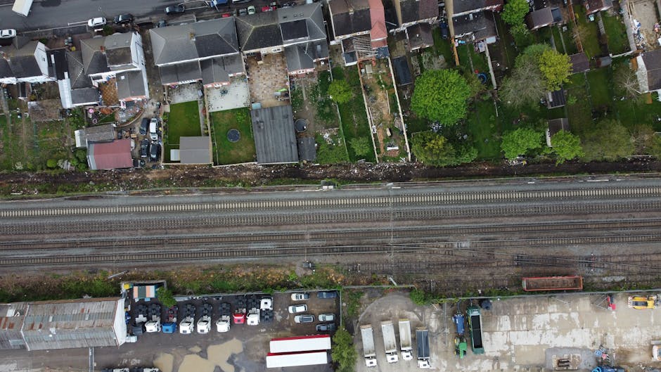 Aerial view of a residential area showing multiple terraced houses with small gardens, some with outdoor patios or lawn areas, separated by narrow pathways. The houses have varied roofing materials, including tiles and flat roofs, and are constructed from brick and wood. Lush green trees and shrubs are visible in several gardens, providing a natural backdrop. Directly below the houses, a railway line with multiple parallel tracks runs horizontally across the image, with a train car visible on one of the tracks. Adjacent to the tracks, there is a parking lot filled with a variety of cars, trucks, and vans, as well as a small warehouse or storage building with a metal roof. An area of open concrete pavement includes additional parked vehicles and some storage containers. The outdoor scene is captured in daylight with natural lighting, illustrating the setting where house removals, furniture transport, and packing and moving activities can take place, with Man with Van Leyton experts regularly assisting with home relocation logistics.