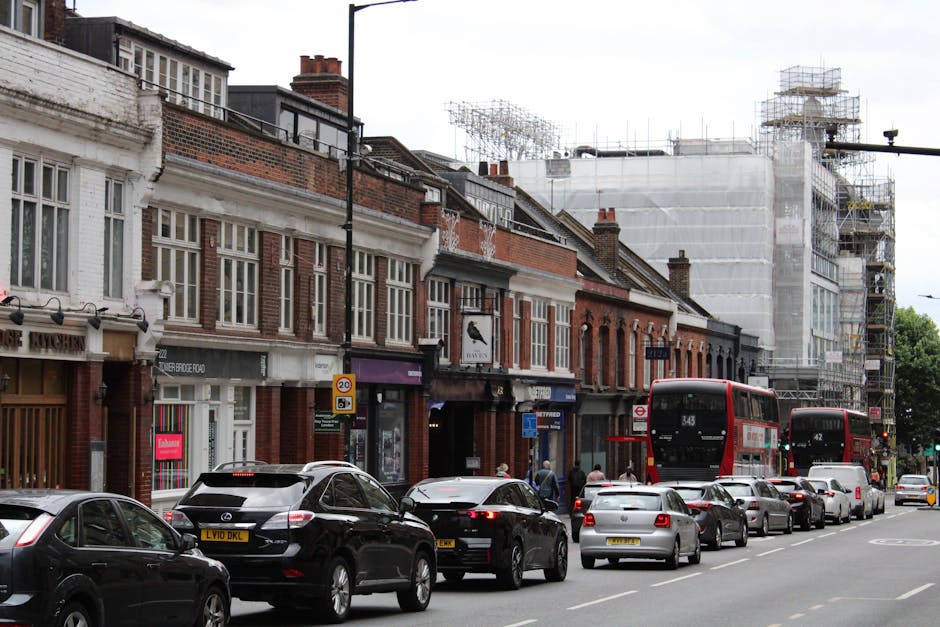 A busy street scene in Leyton showing a row of traditional brick buildings with retail shops on the ground floor, including a shop with a pink sign. Several cars are parked along the curb, with a few in motion, and two red double-decker buses are traveling down the street. On the sidewalk, there are pedestrians walking, and in the background, scaffolding surrounds a building under renovation, indicating ongoing construction work. The environment is daytime with overcast weather, and the scene reflects urban activity typical of home relocation logistics, where vehicles and moving equipment are involved in furniture transport and packing during a moving process. Man with Van Leyton occasionally supports such operations, as suggested by the context.